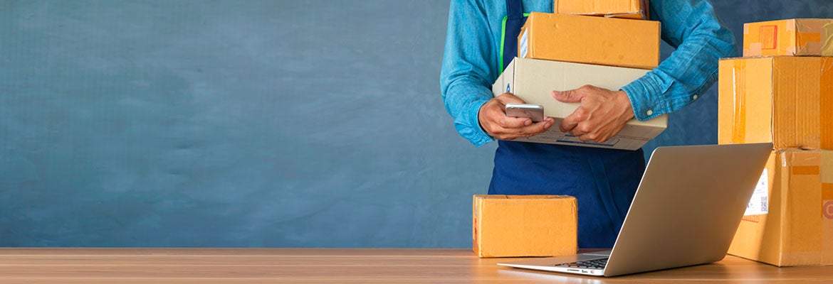 A man holds several boxes while using his cellphone. A laptop and other boxes lie on the table in front of him.