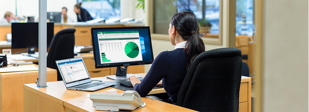 A woman seen from behind sits on her desk at an office and works on some graphs desktop and laptop.