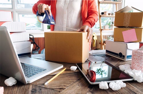 A partial view of a woman taping a cardboard box. In front of her, a laptop, a tablet and other boxes lie on the table.