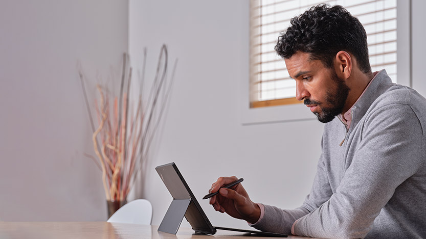 A man sits at a table while working on a Microsoft tablet with a Surface Pen.