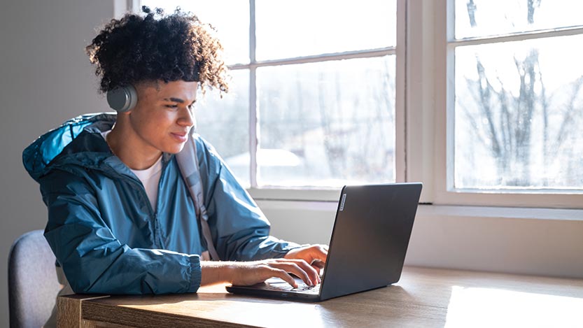 A man sits at a table by a window and works on his laptop computer while wearing headphones.