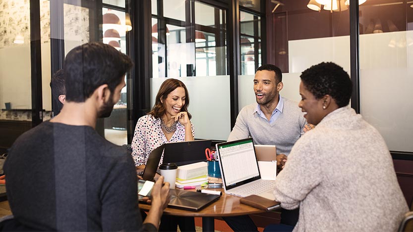 Four people sit at a round table and laugh while working on laptop computers.