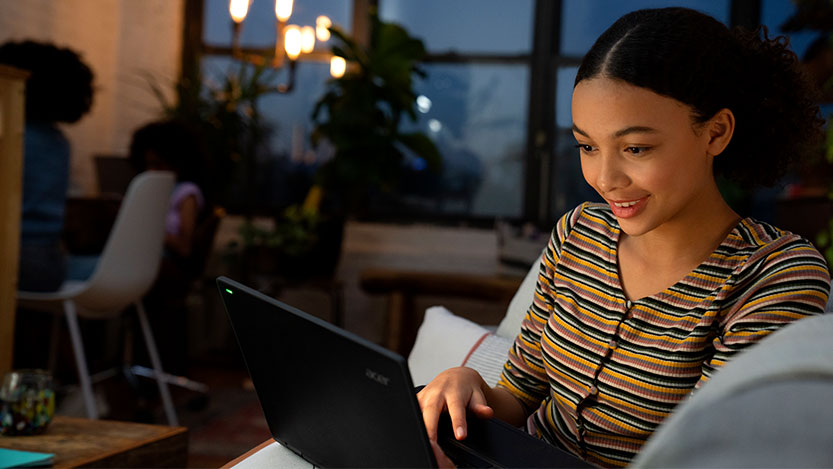 A young woman sits on a couch in a living room and smiles while using a laptop.