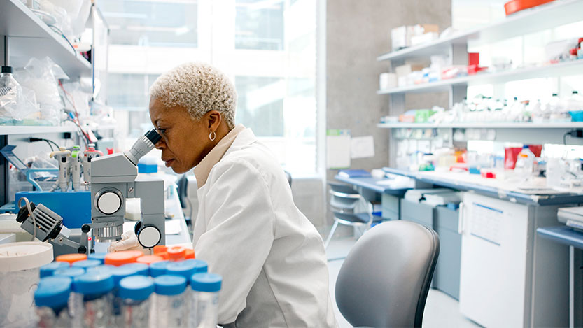 A scientist sits in a lab with a white coat while using a microscope.