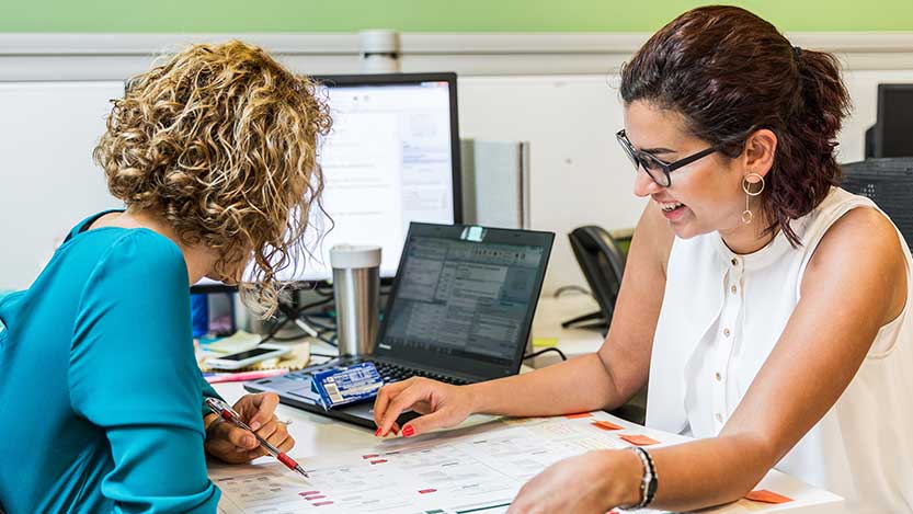 Two women working with some handouts in a desk next to a laptop in an office.