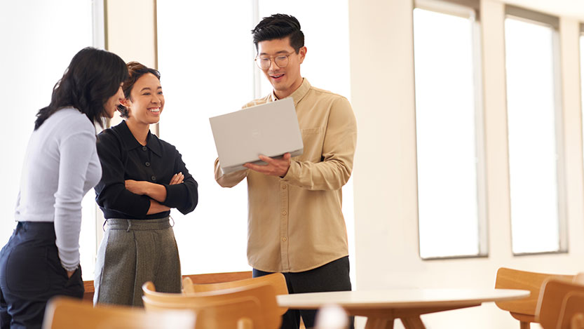 Three people standing in a conference room stare at a laptop computer help up in the air and smile.