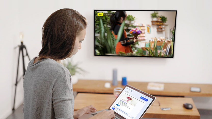 A young woman checking a Video ad on her personal laptop computer while a CTV ad is running on her TV.