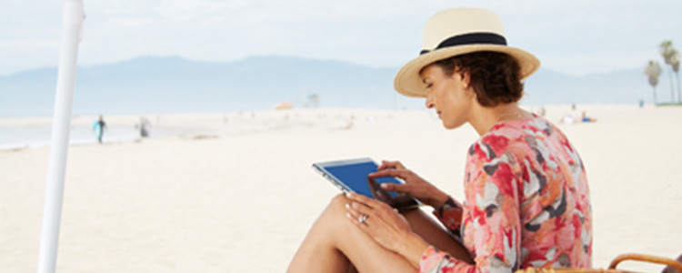 A woman with a hat uses a tablet while sitting on the beach.